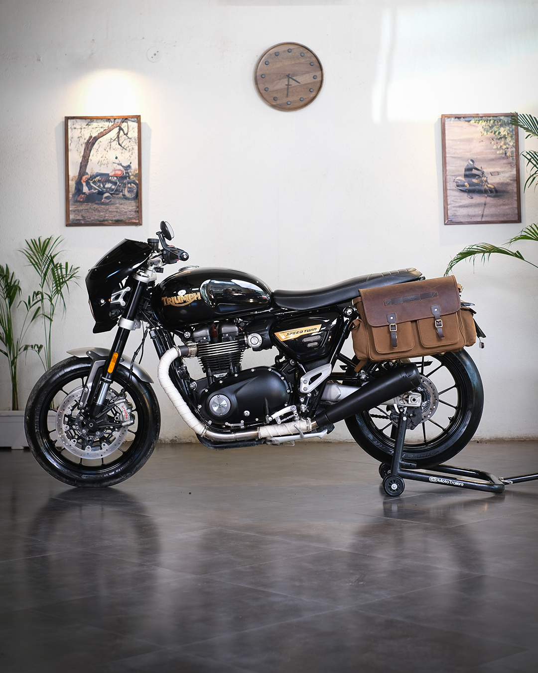 Black motorcycle with a brown bag on a stand in a room with plants and framed pictures.