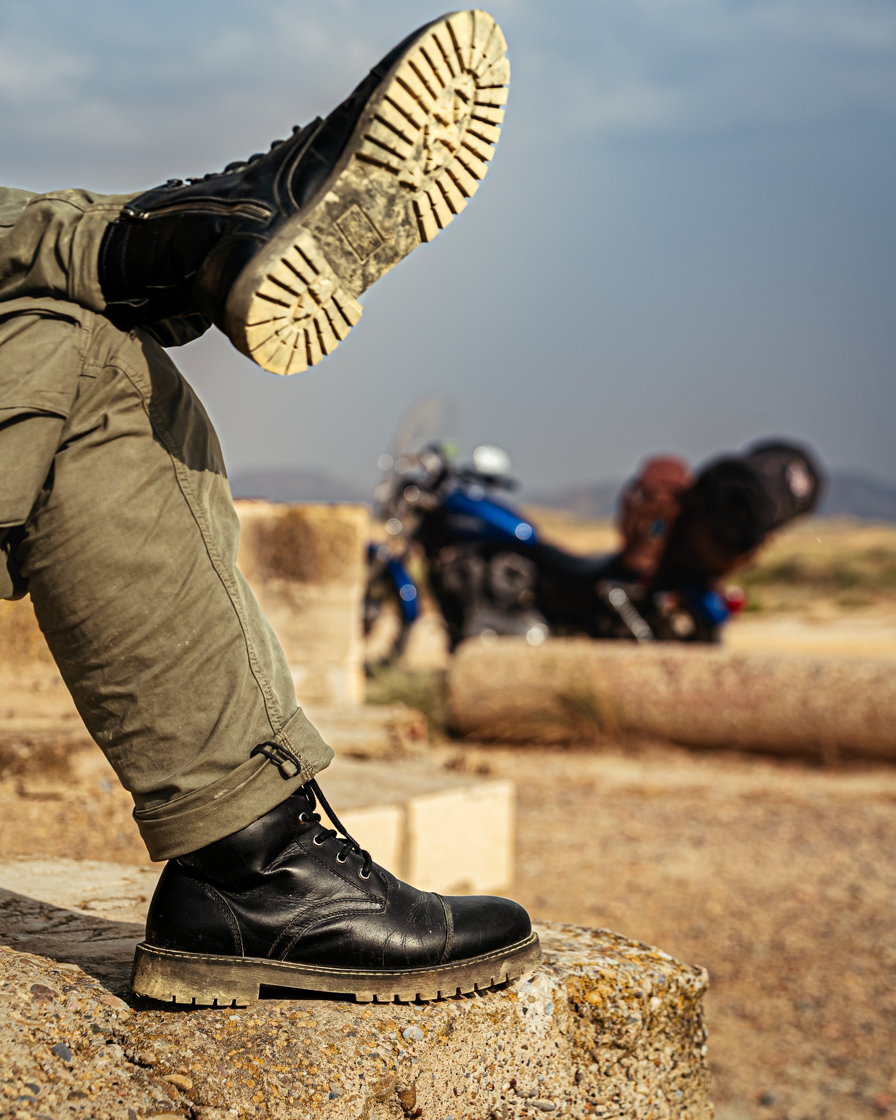 Person wearing black leather boots with a motorcycle in the background