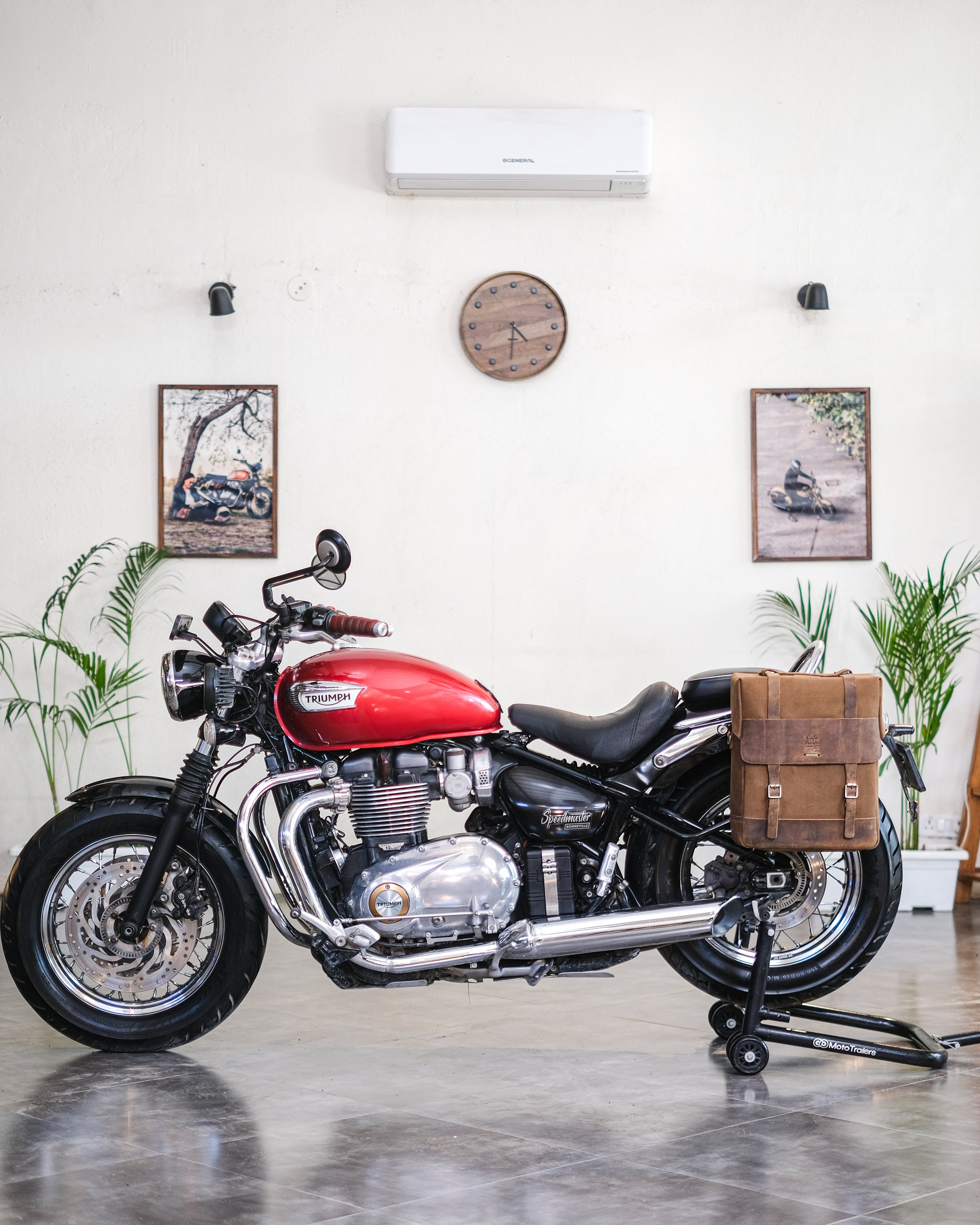 Red motorcycle with a brown leather bag in a room with plants and a clock on the wall.