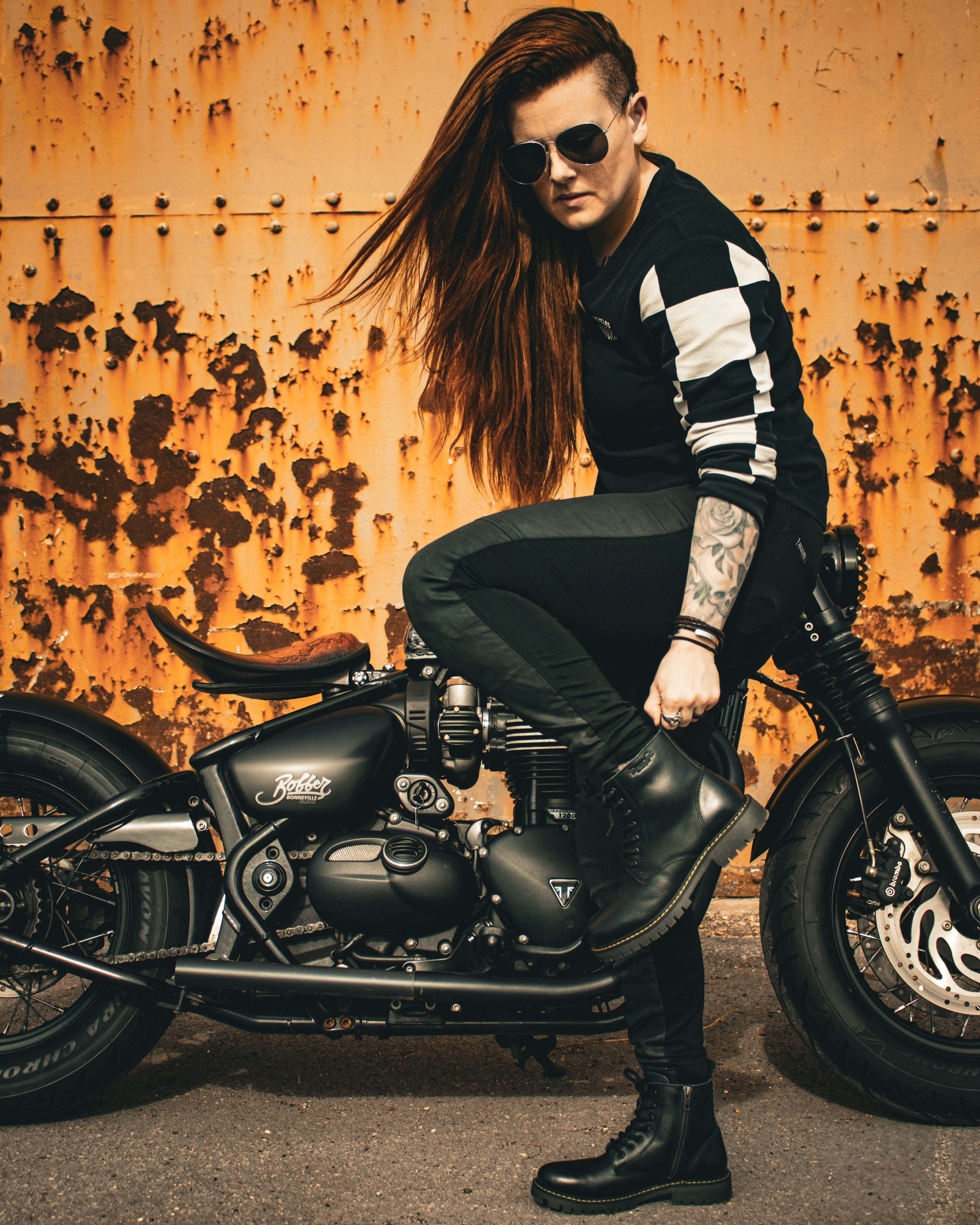 Person with tattoos and sunglasses sitting on a motorcycle against a rusty wall.