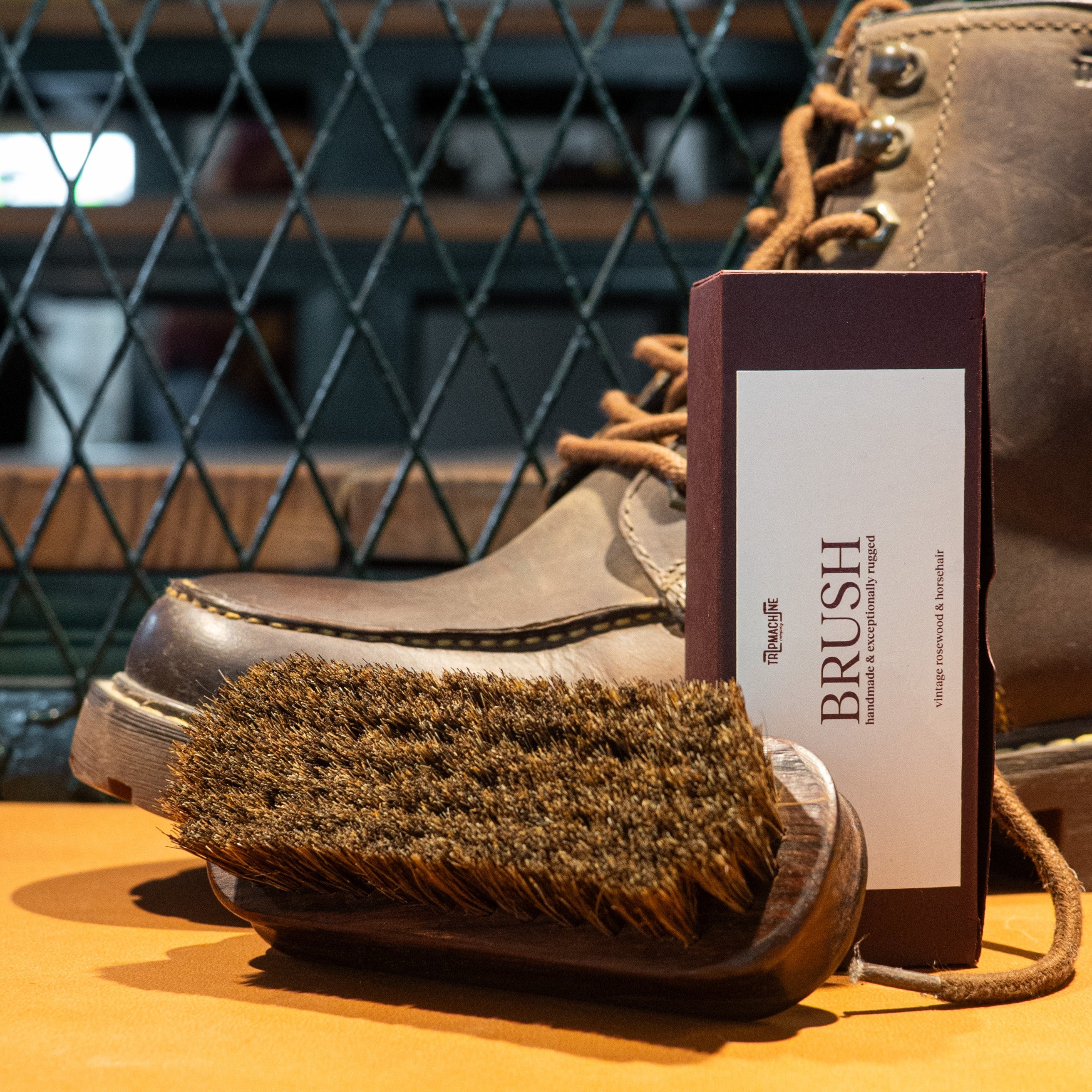Brown leather boot with a brush and care card on an orange surface, against a metal grid background.