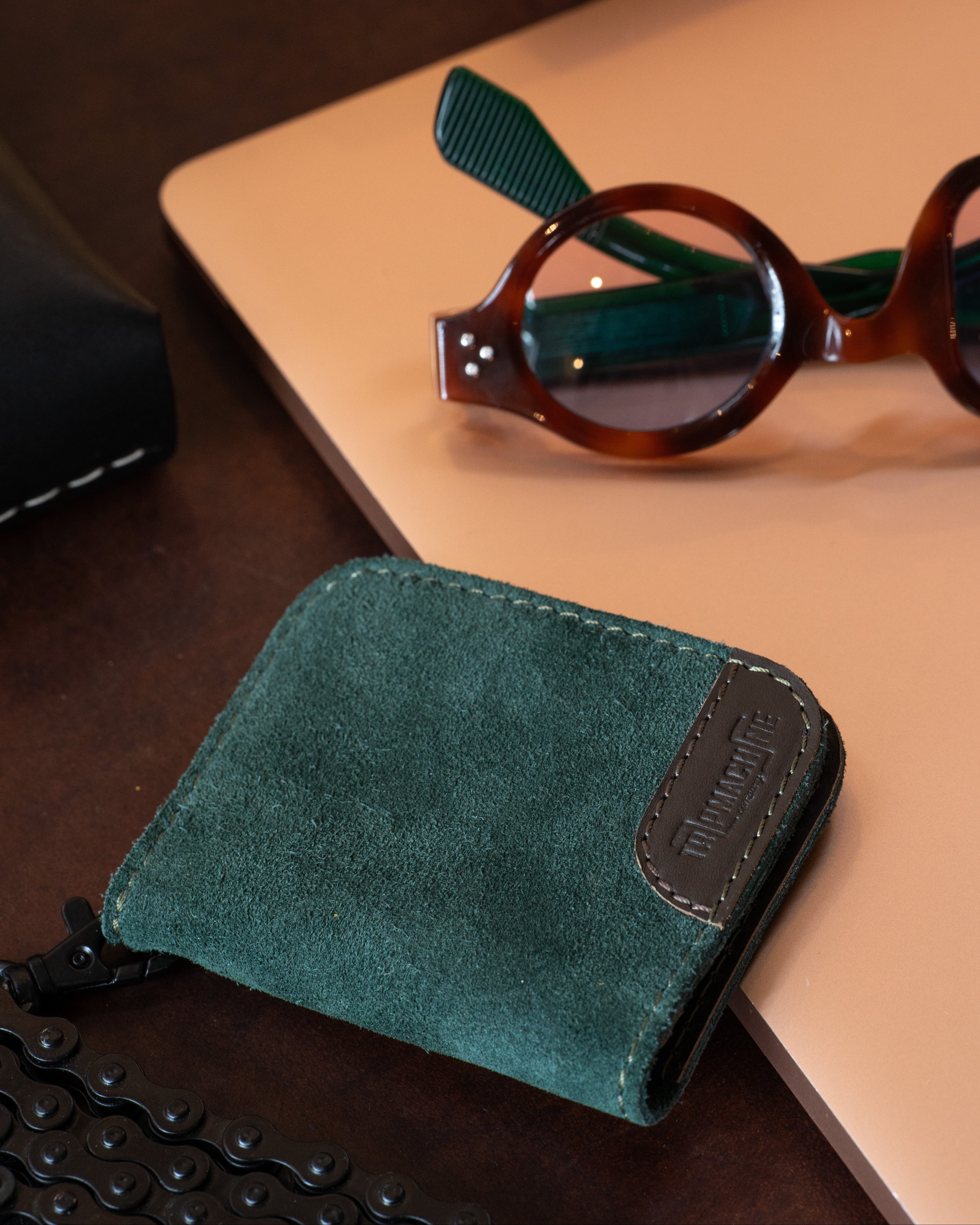 A dark green suede bifold wallet with a brown leather patch and debossed logo, styled on a beige laptop next to tortoiseshell glasses on a dark wooden desk.