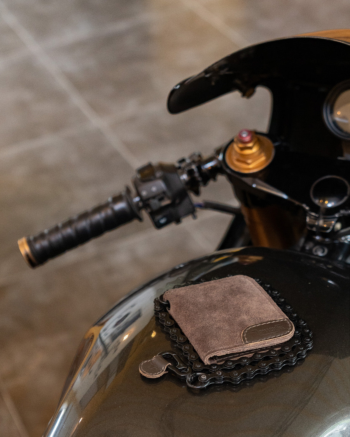 A brown leather bifold wallet with a chain strap detail, resting on a motorcycle tank next to a black chain.