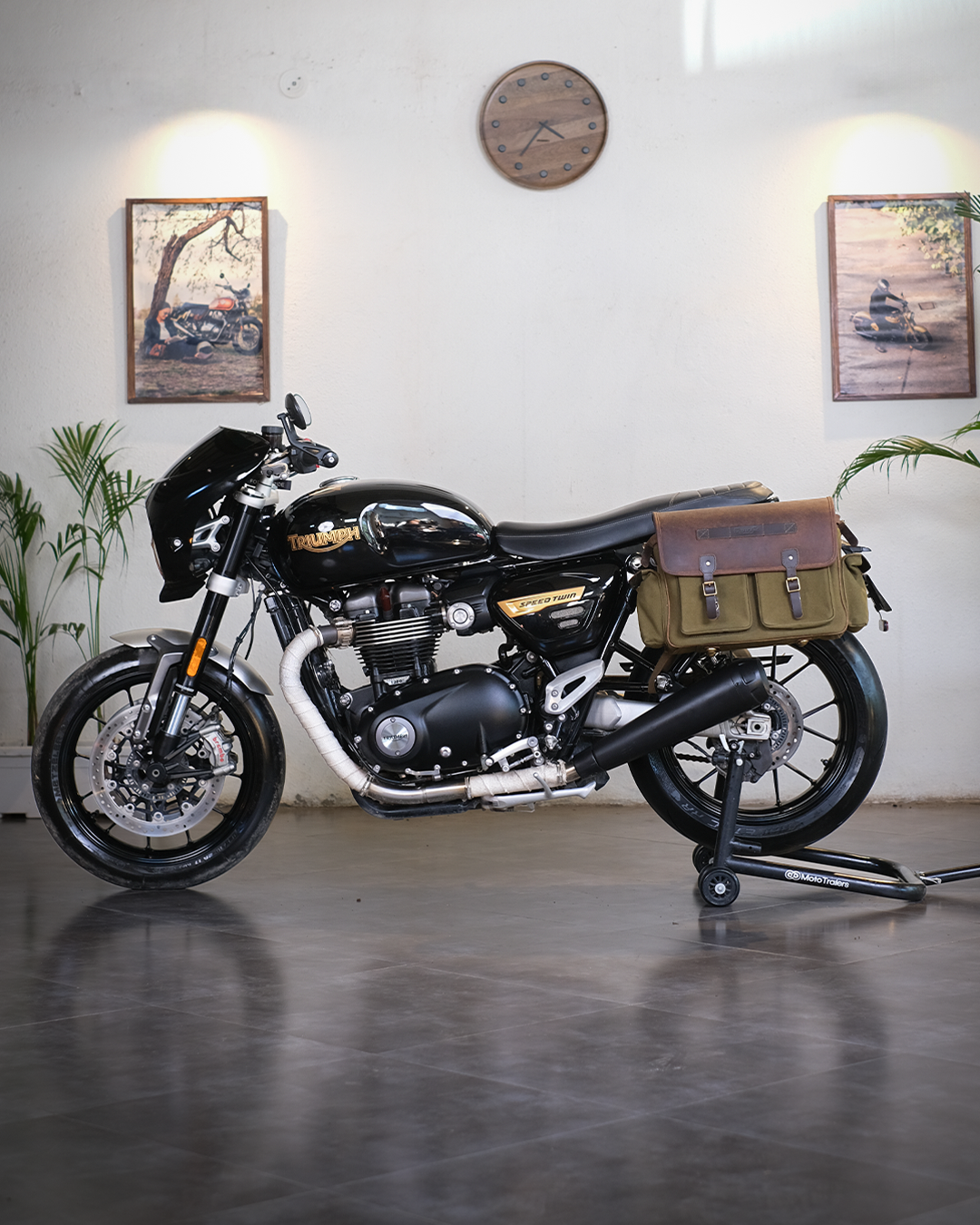 Black motorcycle with a brown bag on a stand in a room with plants and framed pictures on the wall.