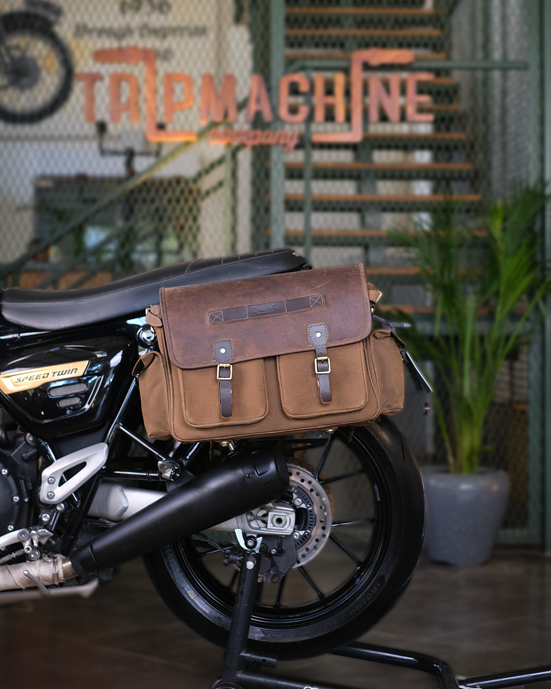 Motorcycle with a brown leather bag attached, set against a backdrop with 'Tailpin Machine' branding.