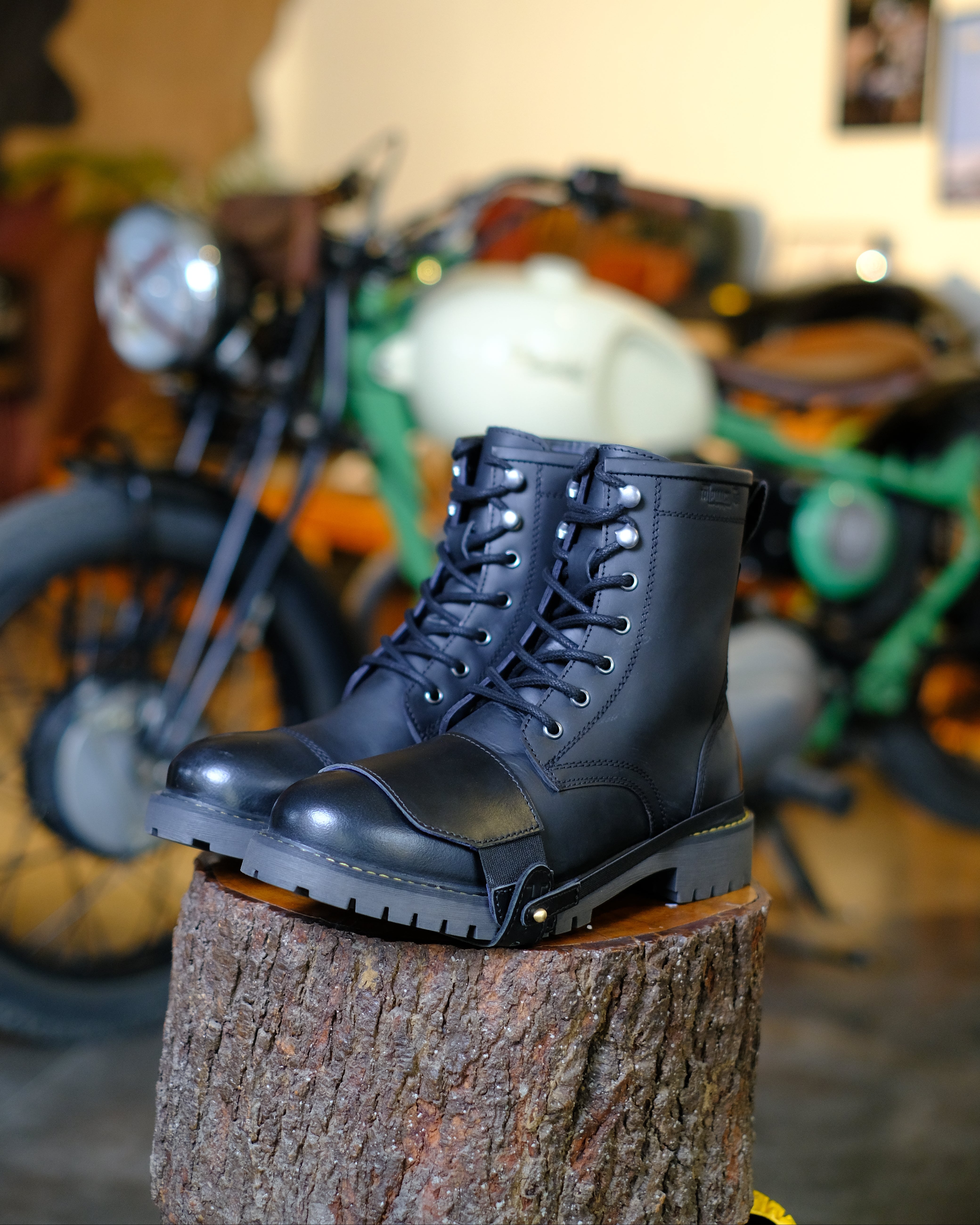 Black leather boots with black shoe protector on a wooden stump with motorcycles in the background