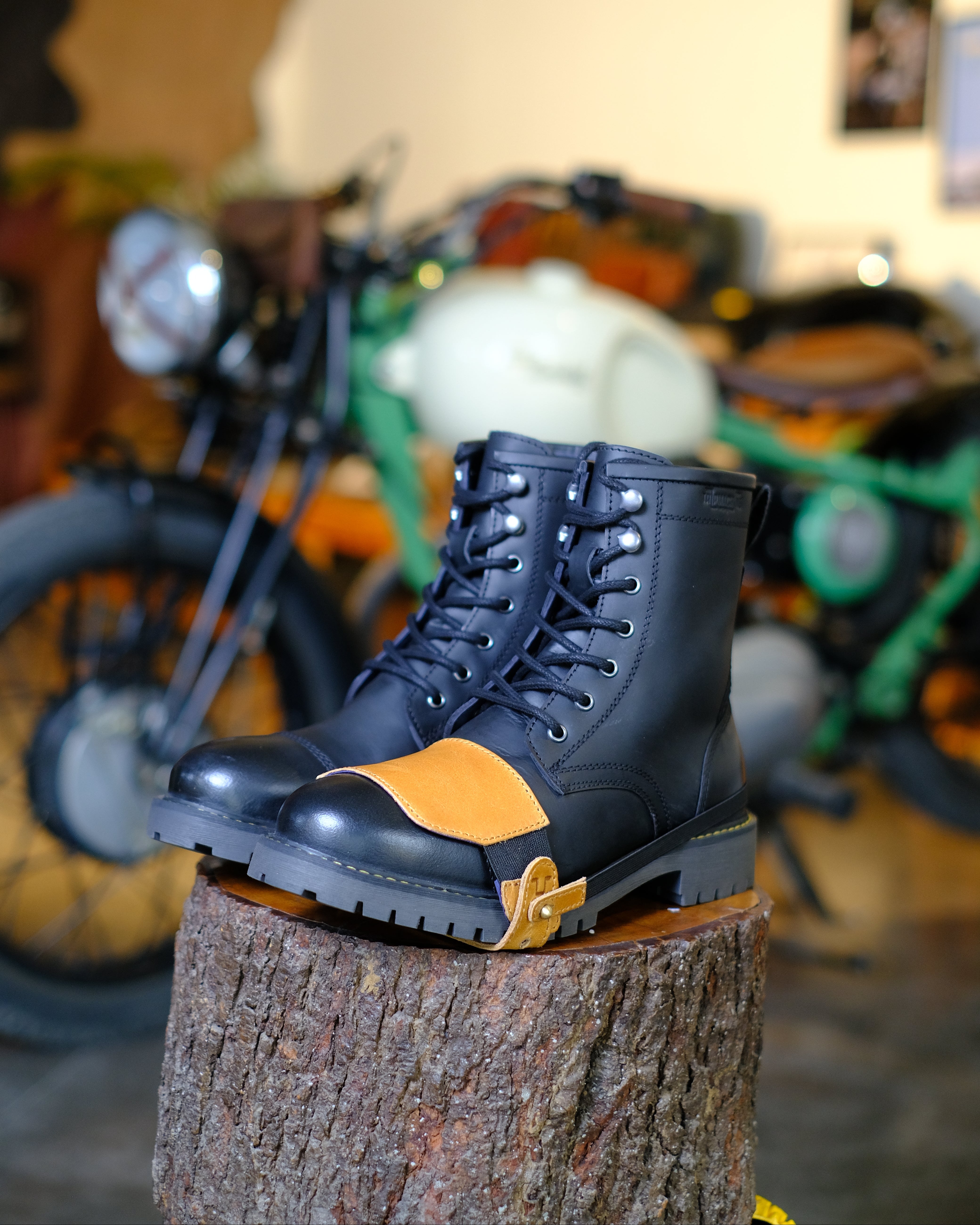 Black boots with tan shoe protector on a wooden block in front of a motorcycle.