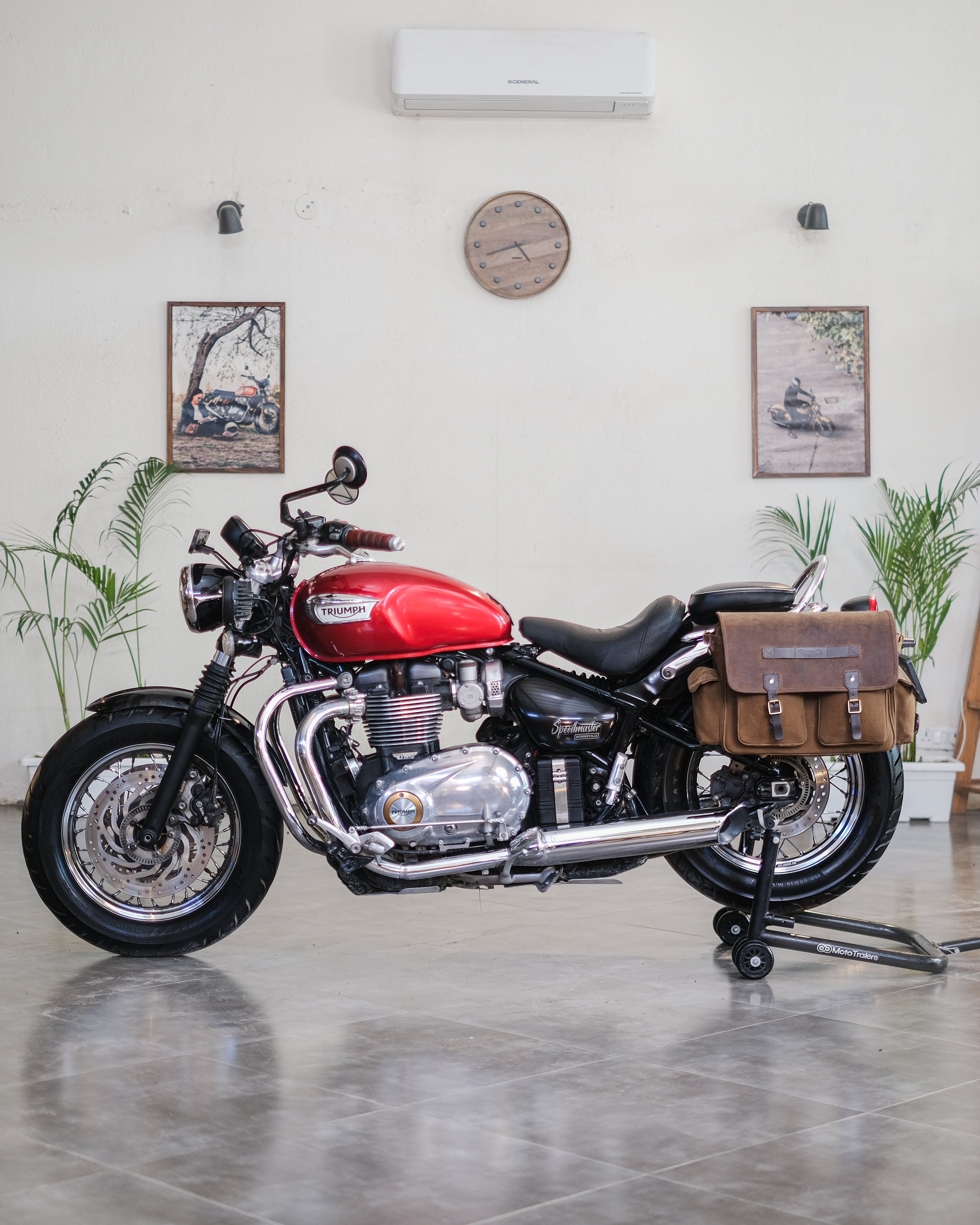 Red motorcycle with a brown saddlebag in a room with plants and framed pictures on the wall.