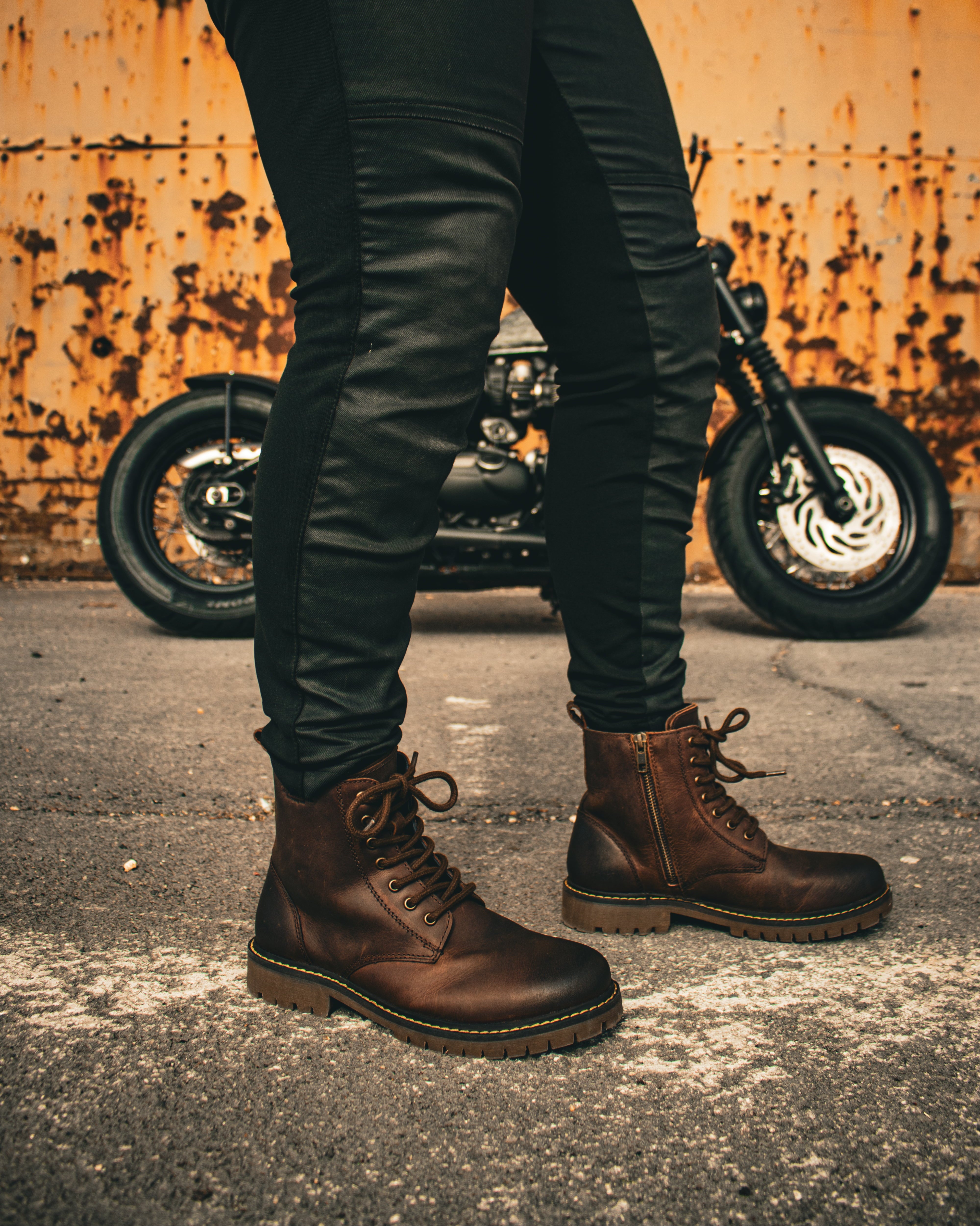 Person wearing dark leather boots and pants standing next to a motorcycle with a rusty wall background