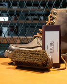 Brown leather boot with a brush and care card on an orange surface, against a metal grid background.