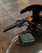 A green suede bifold wallet with a brown leather patch and a small metal keychain, resting on a motorcycle chain in front of a black motorcycle tank.
