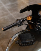 A close-up of a black leather wallet mounted on a black motorcycle chain guard, set against the glossy black fuel tank of a motorcycle.