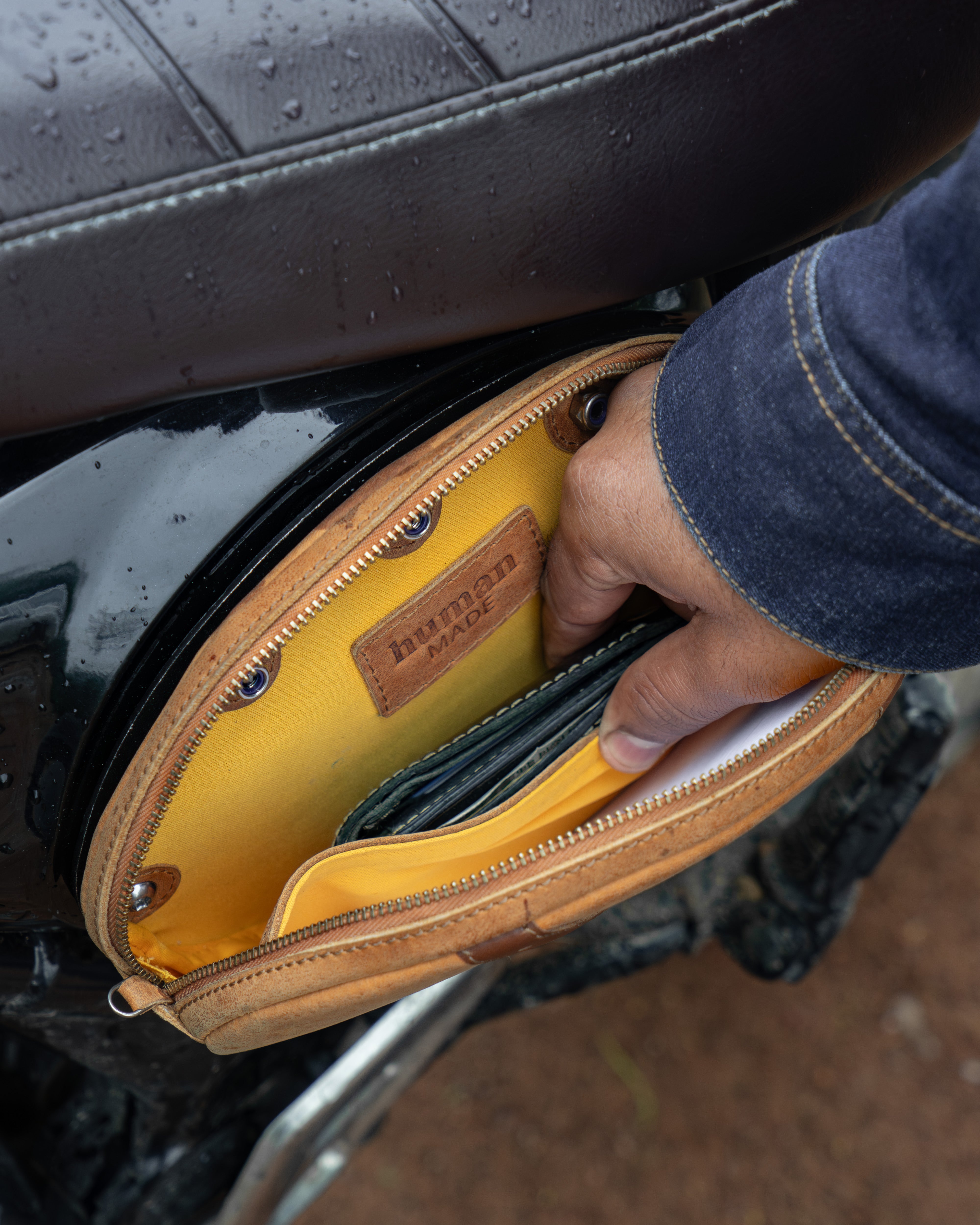 Hand opening a yellow leather bag attached to a motorcycle seat.