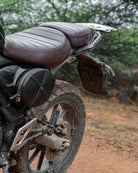 Close-up of a motorcycle seat and rear wheel on a dirt road with trees in the background
