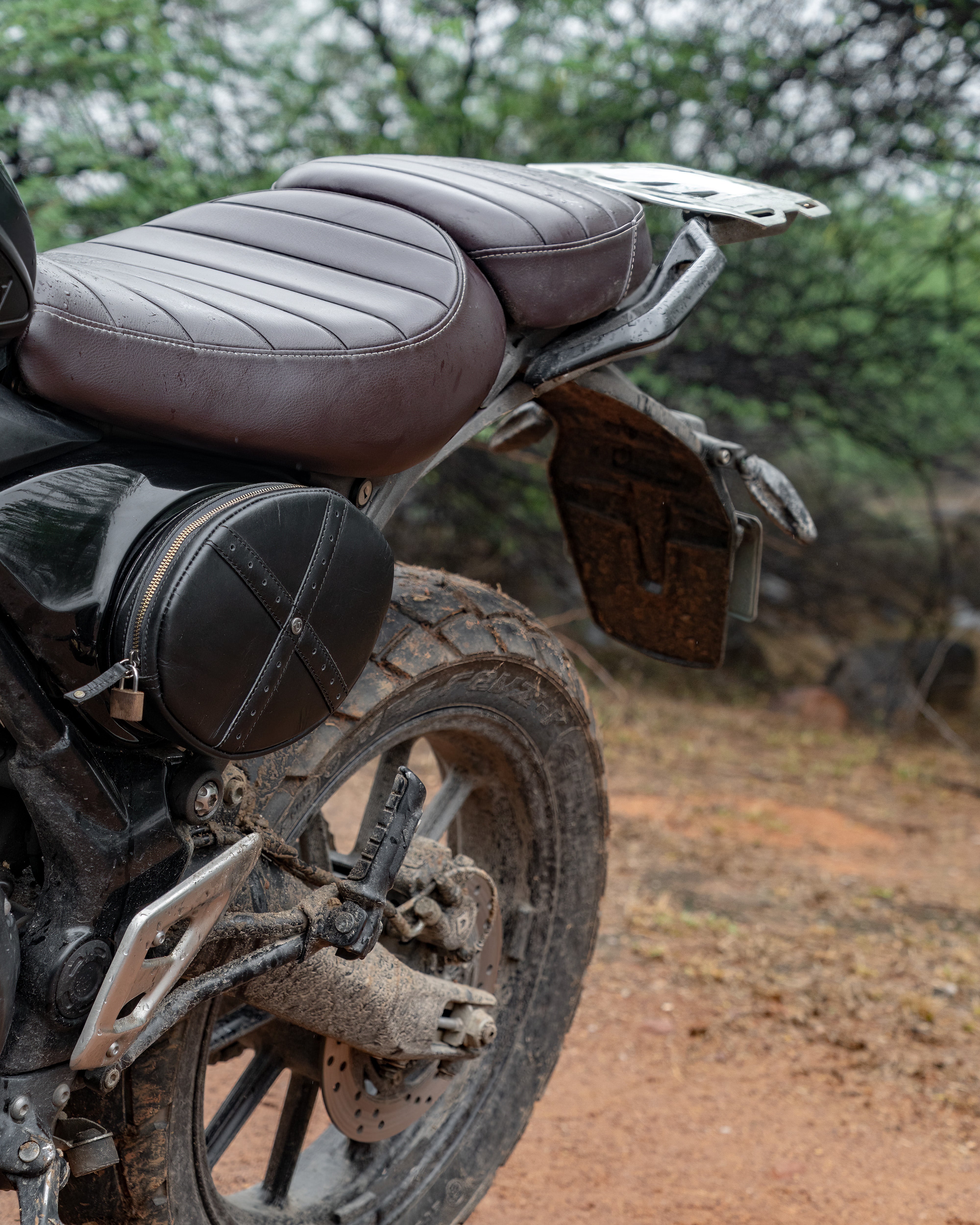 Close-up of a motorcycle seat and rear wheel on a dirt road with trees in the background