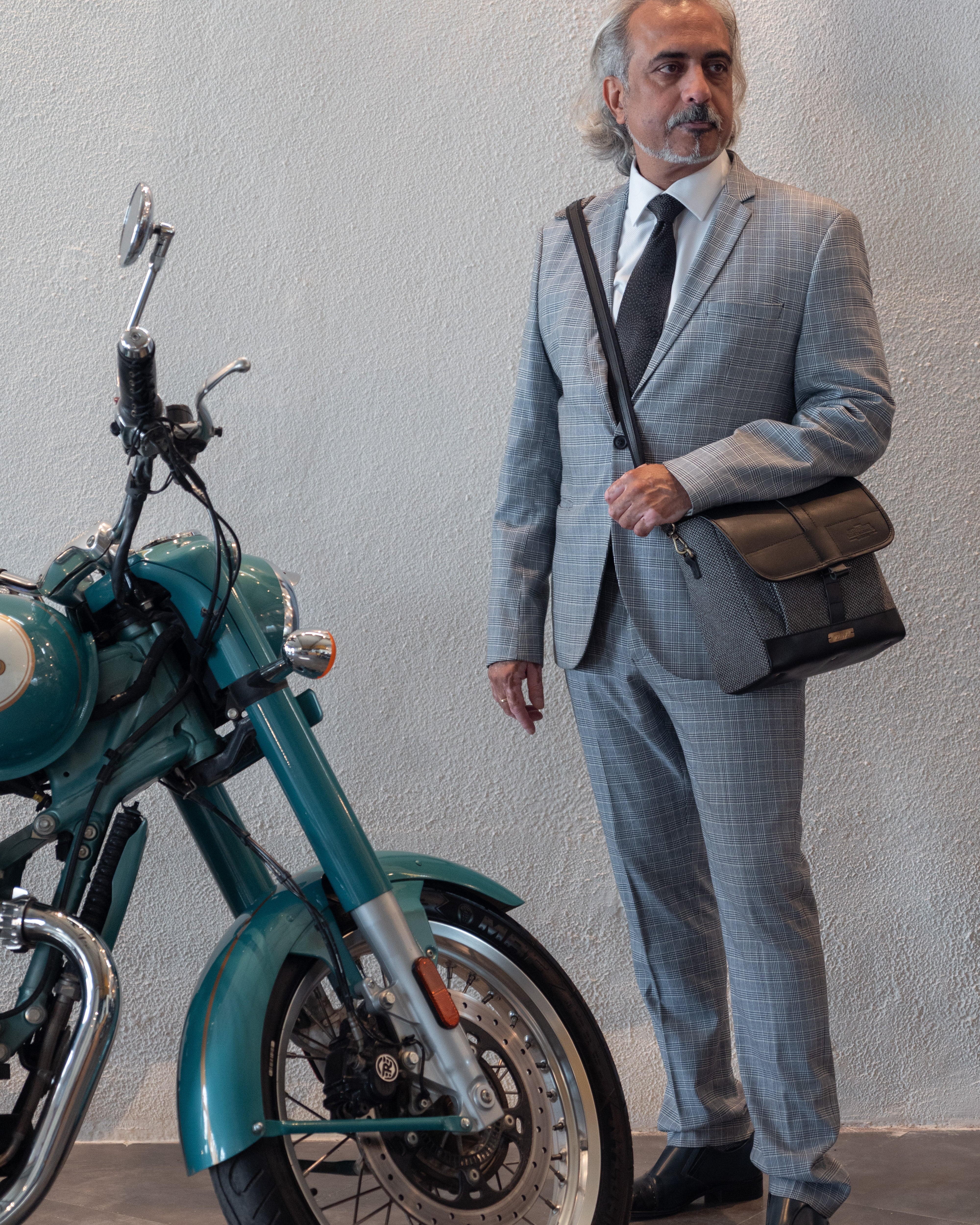 Man in a gray suit standing next to a teal motorcycle against a plain background
