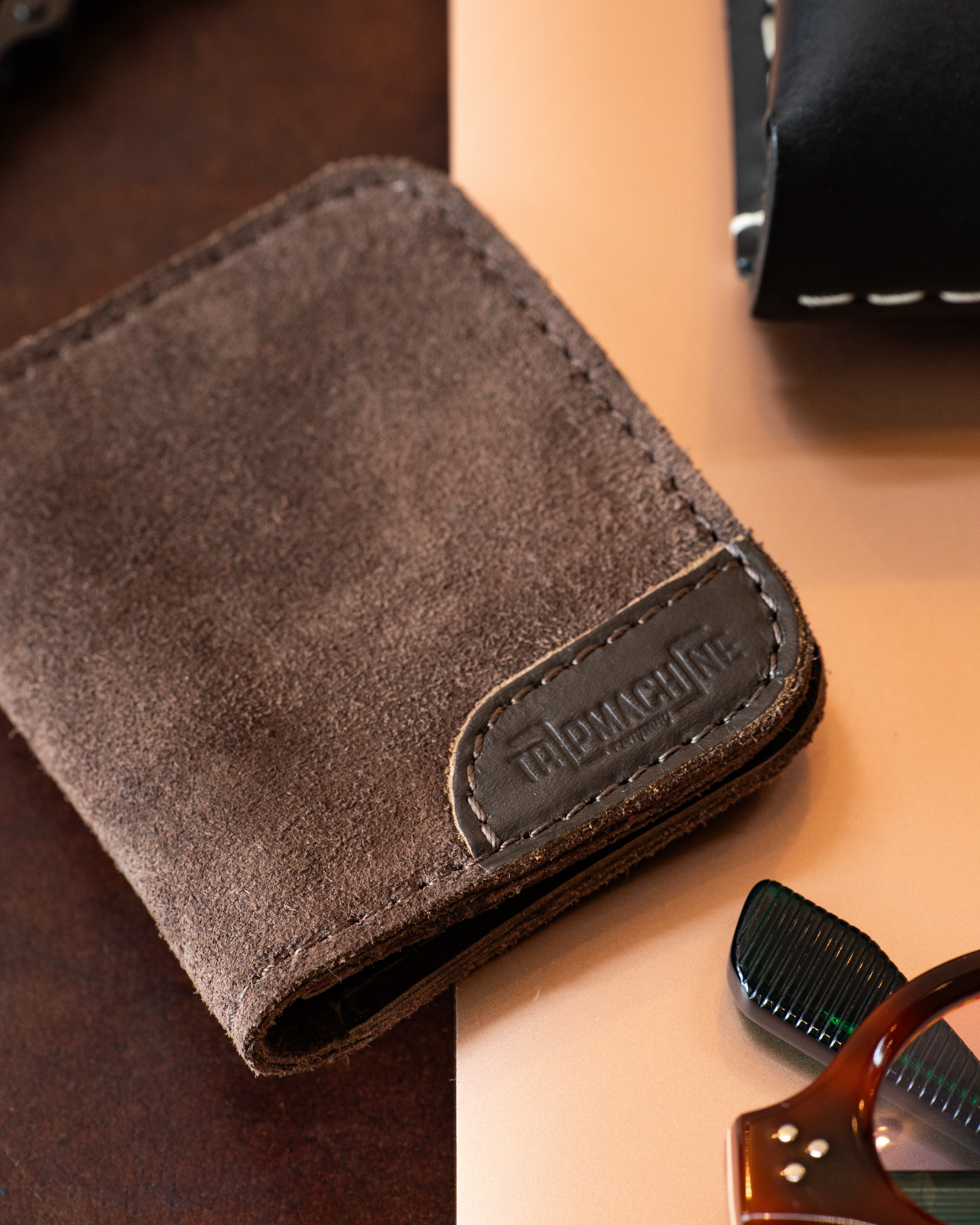 A brown suede bifold wallet with a stitched leather brand patch reading 'TRIP MACHINE' on the front, styled on a split dark wood and peach background with partial props visible.